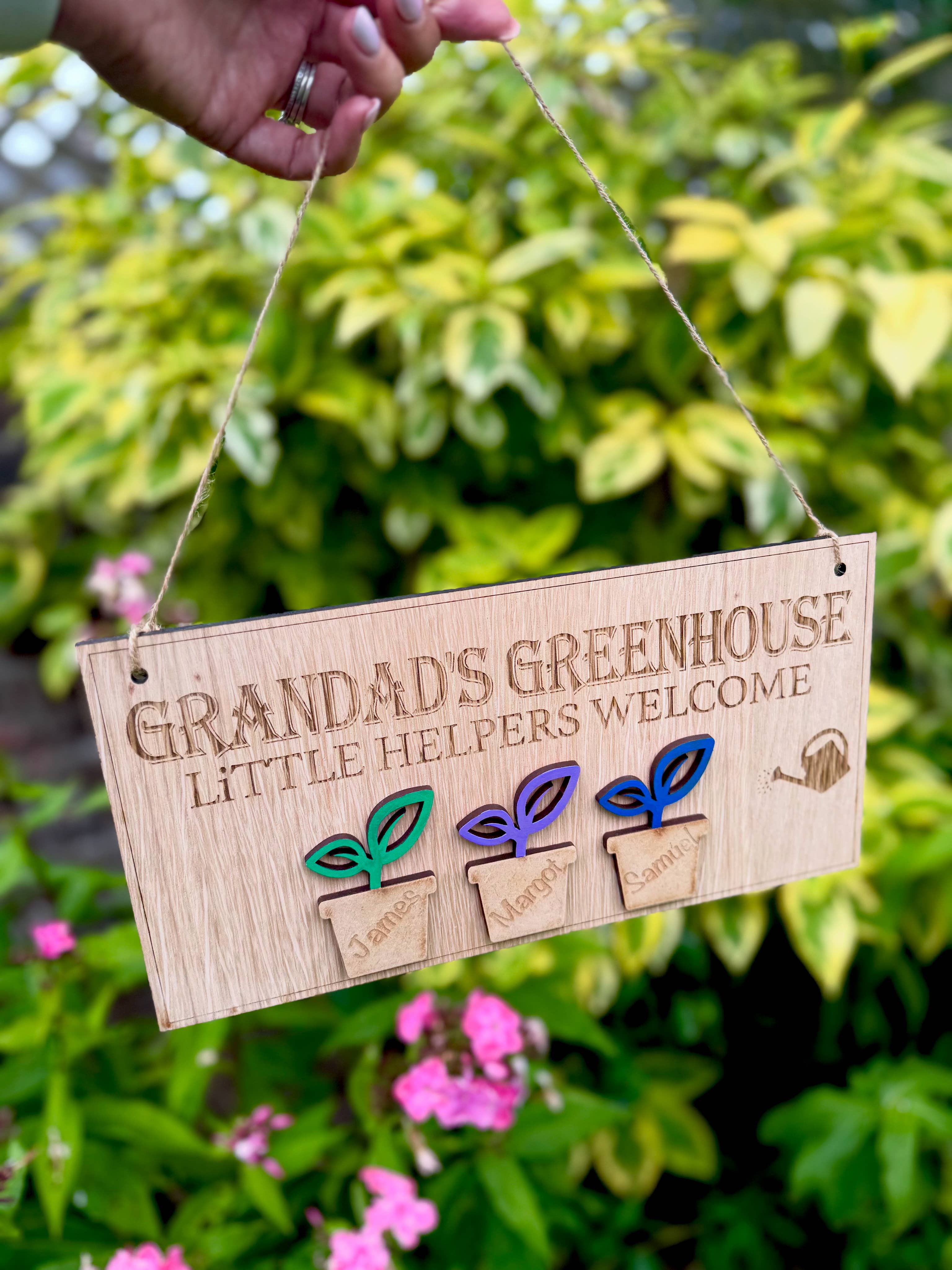 Personalised wooden sign reading โGrandadโs Greenhouse โ Little Helpers Welcomeโ with a plant pot labelled with childrens names, hanging inside a bright sunny garden.